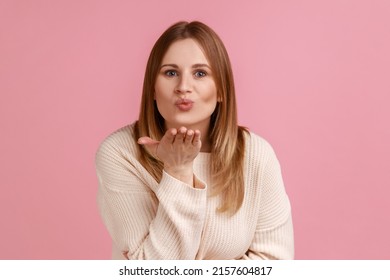 Portrait of romantic blond woman sending air kiss over palms, expressing fondness, romantic feelings and flirt, wearing white sweater. Indoor studio shot isolated on pink background.