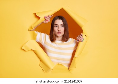Portrait of romantic lovely adorable woman stands in torn paper hole, posing with pout lips and sending air kisses, looking through breakthrough of yellow background.