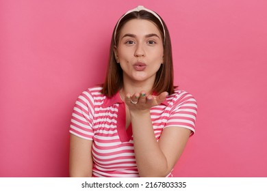 Portrait of romantic lovely Caucasian woman wearing striped T-shirt and hair band, sending air kissing to her boyfriend, posing isolated over pink background.