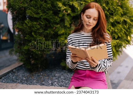 Portrait of romantic redhead female sitting on bench in city park and reading paper book. Closeup front view of smiling young woman female student reading textbook in nature.