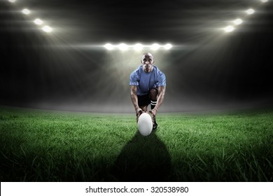 Portrait of rugby player holding ball while kneeling against rugby stadium - Powered by Shutterstock