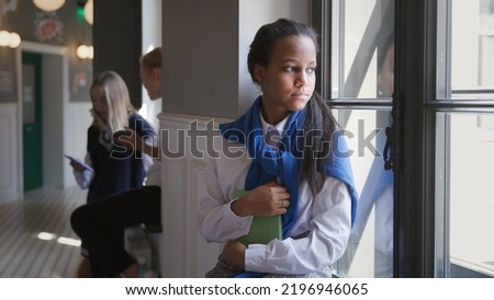 Portrait of sad African-American teen student sitting on window sill in corridor. Upset schoolgirl having problems with education or failing examination looking out of window