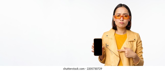 Portrait Of Sad Asian Woman In Sunglasses, Pointing Finger At Mobile Phone App Interface, Showing Smartphone Application, Standing Over White Background