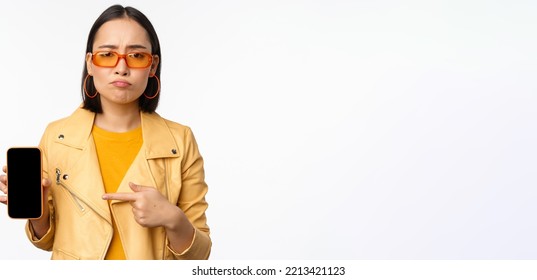 Portrait Of Sad Asian Woman In Sunglasses, Pointing Finger At Mobile Phone App Interface, Showing Smartphone Application, Standing Over White Background