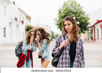 Portrait of sad beautiful teen girl in checked shirt with braids looking at camera against two friends in sunglasses and denim clothes gossiping about her in the street. Unfocused.