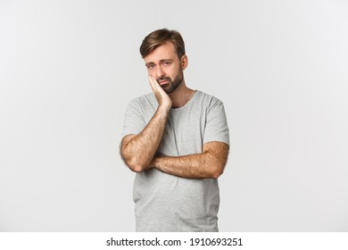 Portrait Of Sad And Bored Bearded Man In Gray T-shirt, Leaning On Palm And Looking Reluctant, Standing Over White Background