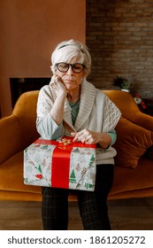 Portrait Of Sad And Bored Elder Woman Sitting Alone On A Sofa With Wrapped Gift. No Family Visit Her On Christmas. Sadness During Coronavirus Pandemic. Covid Quarantine, Lockdown.