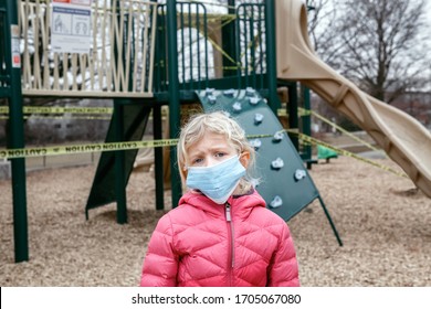 Portrait Of Sad Caucasian Girl In Face Mask On Closed Playground Outdoor. Kids Play Area Locked With Yellow Caution Tape In Toronto Canada. Coronavirus Social Distance Quarantine.