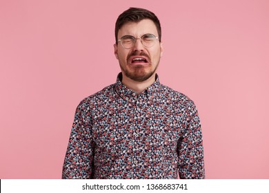 Portrait of a sad crying bearded man in glasses, wearing in colorful shirt, looks unhappy and upset. Isolated over pink background, people and emotions concept.