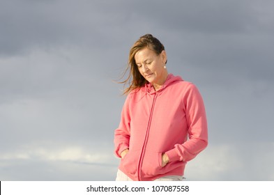 Portrait Of Sad And Depressed Looking Mature Woman In Pink Sweater, At Beach, Isolated With Storm Clouds As Background And Copy Space.