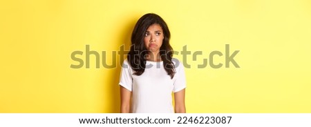 Portrait of sad and disappointed african-american woman, sulking and frowning as looking at upper left corner, see something upsetting, standing over yellow background.