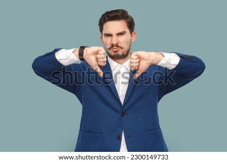 Portrait of sad displeased handsome man with mustache standing and showing dislike gesture, disagree, wearing official style suit. Indoor studio shot isolated on light blue background.
