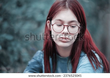 Portrait of a sad girl with glasses. A thoughtful red-haired teenage girl in a blue T-shirt with downcast eyes.