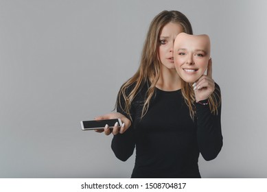 Portrait of a sad girl with a smartphone in one hand and a smiling mask in the other hand, with which she covers her face, as a symbol of the inconsistency of the image of a person in social networks