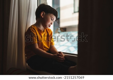 Portrait of sad and gloomy little boy sulking from disappointment, sitting on windowsill upset and look at window. Kid curves lower lip, he wear yellow t-shirt at home. 