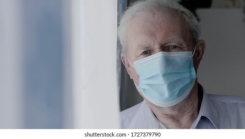 Portrait of sad grandfather in medical mask looks at camera anxiously during self-isolation at home. 4K