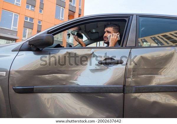 Portrait of sad handsome\
bearded man sitting in dented car and talking cell phone, calling\
to insurance inspection or asking somebody to help, road accident.\
Outdoor shot.