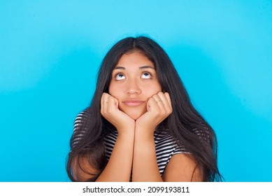 Portrait Of Sad Hispanic Girl Wearing Striped T-shirt Standing Over Blue Background Hands Face Look Empty Space