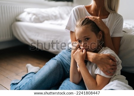 Portrait of sad little girl looking at camera and loving caring mother comforting offended afraid child daughter, showing love and care, expressing support, hugging and stroking hair sitting on floor.