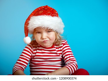 Portrait Of Sad Little Girl In Red Santa Claus Hat. Little Child Waiting For Christmas Presents. Funny Kid Against Blue Wall At Home In Christmas Night. Happy Xmas And New Year, Winter Holiday