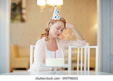 Portrait Of Sad  Lonely Mature Woman Sitting Alone At Birthday Table With Cake, Wearing Party Hat And Waiting For Guests