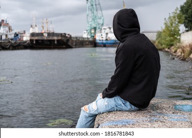 Portrait Of A Sad Lonely Young Guy, A Teenager In A Black Hoodie On The River Bank Near The Port. Problems, Loneliness, Adolescent Psychology, Concept.