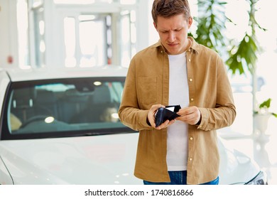 Portrait Of Sad Man Having No Money To Buy New Car. Man Stand In Dealership Looking At Purse, White Auto In The Background. Crisis In Country Hits The Wallet