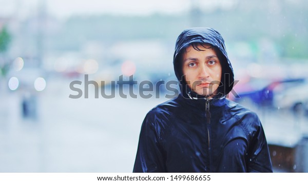 Portrait of a sad man in a\
raincoat and a hood in the rain. Bad stormy rainy weather sorrow\
concept.