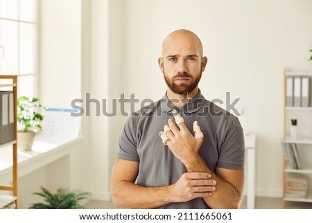 Portrait of sad man who has hurt his fingers and is now wearing adjustable finger splint braces. Caucasian bald young bearded man who injured four fingers on his left hand looks at camera.