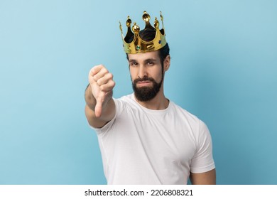 Portrait Of Sad Serious Man With Beard In White T-shirt And In Gold Crown Standing Looking At Camera With Disappointed Expression, Showing Thumb Down. Indoor Studio Shot Isolated On Blue Background.