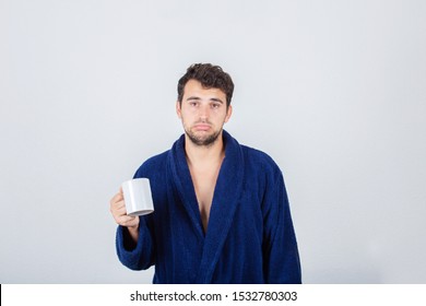 Portrait of sad sleepy young man messy hair, wears blue bathrobe, drinking coffee from his cup isolated on white background.