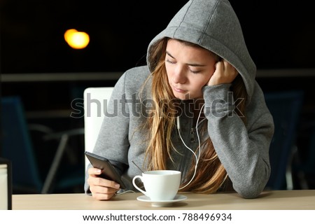 Portrait of a sad teen listening to music with a smart phone and earphones in a coffee shop in the night