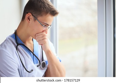 Portrait Of Sad And Unhappy Young Doctor Thinking Near A Window