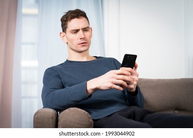 Portrait of sad unpleasant unhappy guy, young frowning man looking at screen of his cell mobile phone, using smartphone with negative face, sitting at home in living room on sofa or couch. Bad news