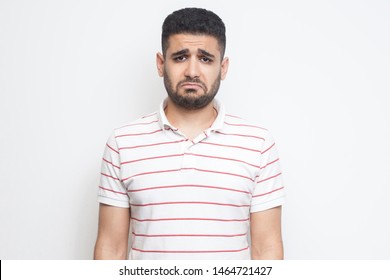 Portrait of sad updet or bored bearded young man in striped t-shirt standing and looking at camera with dissatisfied sadness face. indoor studio shot, isolated on white background.