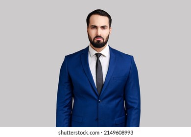 Portrait of sad upset bearded man standing and looking at camera with dissatisfied sadness face, expressing sorrow, wearing official style suit. Indoor studio shot isolated on gray background.