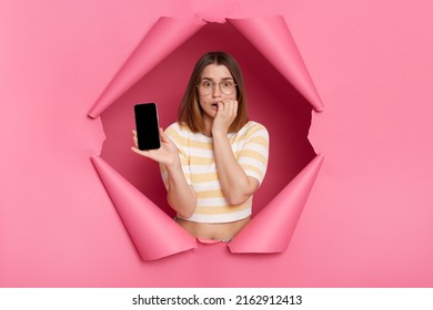 Portrait Of Sad Upset Caucasian Woman Breaks Through Pink Paper Background, Standing Showing Cell Phone With Blank Display With Advertisement Area For Promotional Text.