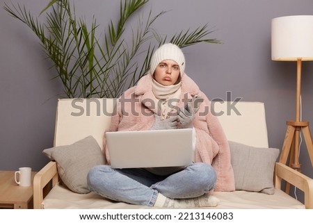Portrait of sad upset frozen woman wearing cap, gloves, scarf and coat sitting in living room, holding smart phone in hands, holds laptop on her knees, posing in cold house.