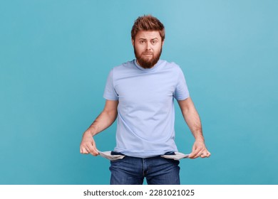 Portrait of sad upset poor bearded man showing empty pockets and looking frustrated about loans and debts, has no money, being jobless. Indoor studio shot isolated on blue background. - Shutterstock ID 2281021025