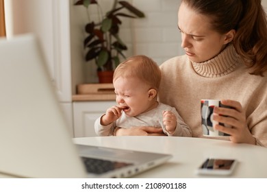 Portrait Of Sad Woman Wearing Beige Casual Style Sweater With Ponytail Hairstyle Sitting At Table With Her Crying Infant Daughter, Holding Cup Of Coffee Or Tea, Trying To Calm Down Baby.