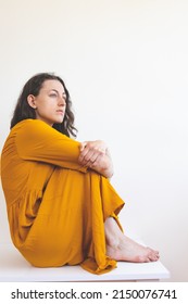 Portrait Of A Sad Woman In A Yellow Dress, Pensive Brunette, Sad Mood, Melancholy, Girl In A White Room Sits On A Table