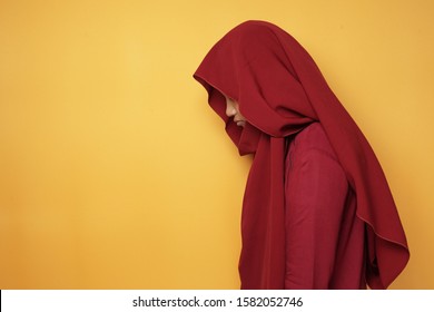 Portrait Of Sad Young Asian Teenage Muslim Girl Wearing Red Hijab Looking Down And Depressed, Side View Profile Standing Against Yellow Background