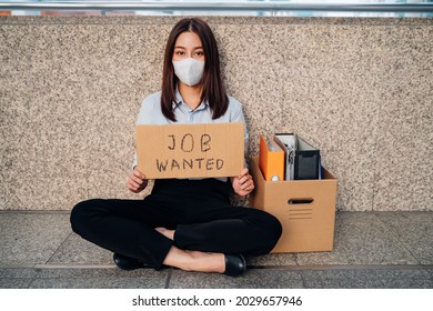 Portrait Of Sad Young Asian Woman With Box Of Items Sitting Alone After Being Laid Off From Job Due To Covid-19 Pandemic Holding A Placard With Job Wanted Text