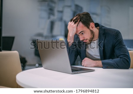 Portrait of a sad young businessman at a working laptop. Young handsome man in a jacket in the interiors of a modern office preoccupied with problems sits at a computer
