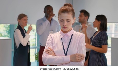 Portrait Of Sad Young Businesswoman Standing Alone At Office Party And Looking At Camera With Colleagues Having Fun And Celebrating On Background