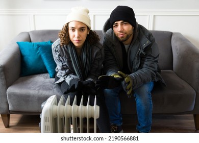 Portrait Of A Sad Young Couple Looking At The Camera While Trying To Stay Warm With A Heater During A Cold Winter At Home