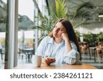 Portrait of sad young redhead woman in a coffee shop looking at smart phone. A lonely unhappy 30s girl siting in a cafe. Bad feelings stress, anxiety, grief, emotions.