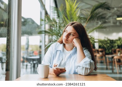 Portrait of sad young redhead woman in a coffee shop looking at smart phone. A lonely unhappy 30s girl siting in a cafe. Bad feelings stress, anxiety, grief, emotions. - Powered by Shutterstock