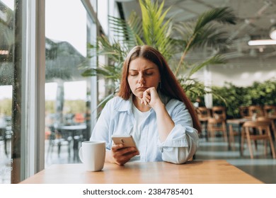 Portrait of sad young redhead woman in a coffee shop looking at smart phone. A lonely unhappy 30s girl siting in a cafe. Bad feelings stress, anxiety, grief, emotions. - Powered by Shutterstock