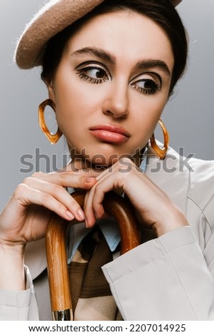 portrait of sad young woman in beret and trench coat posing with umbrella handle isolated on grey